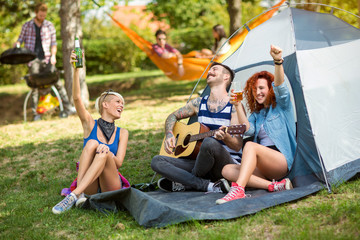 Cheerful youth drinks beer and play guitar in camp