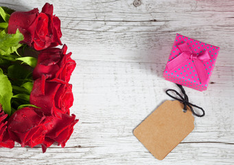 Red roses and small pink gift box with empty tag on white rustic wooden table. Top view with copy space.