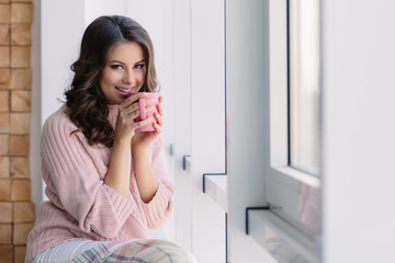 Pretty young woman in the pink sweater sitting nestled plaid and drinking coffee on the windowsill near the window