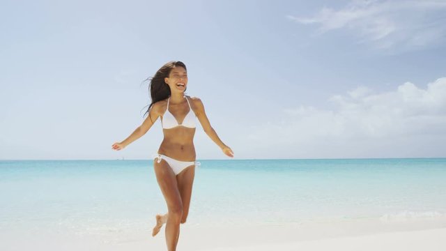 Beach bikini woman carefree running in freedom fun. Joyful happy Asian girl relaxing showing joy and happiness in slim body for weight loss diet concept on perfect white sand. Throwing beach hat.