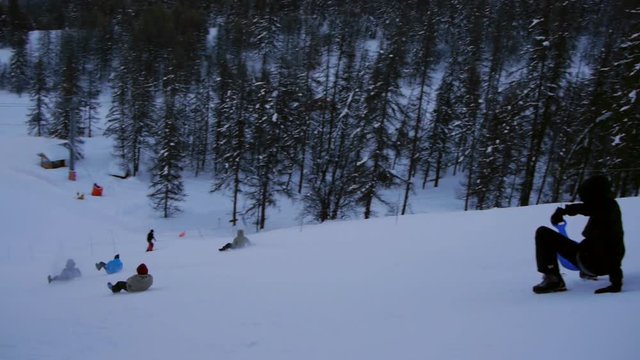 Playful group of teenagers takes a sledding ride down the ski slope and have fun times after skiing day. At the end tilt up to beautiful mountain range at dusk.
