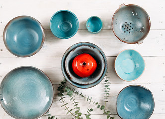 Many turquoise clay empty plates and red decorative ceramic pomegranate  on white wooden background. Flat lay on set of trendy  empty plates and color decor. Organic food, pottery, decoration concept