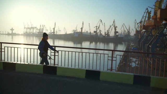 young man holding a skateboard walking along the sea port at dawn slow motion