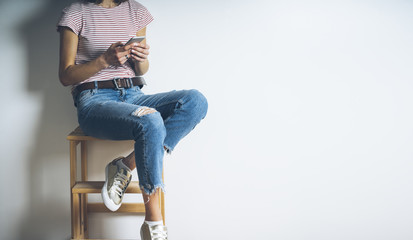 Close-up image of hipster girl sitting on a char and using modern smart phone, blank white...