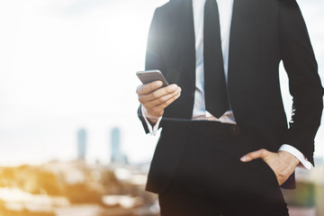 Close-up of young professional businessman wearing modern suit and using smartphone outdoors, blurred background with copy space for your content or design