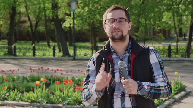 Male reporter with a microphone tells news camera. It should be in the spring park on a background of trees and flowers