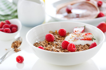 Homemade oat meal granola or muesli with fresh summer fruits – raspberry and strawberry with yogurt in a white bowl on a table for breakfast, closeup.