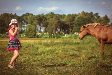 Beautiful scene. young girl on a meadow. Brown horse in the background blurred. Sunny summer day.  childhood, leisure, friendship and people concept.  Children playing outdoors.  Happy family. series