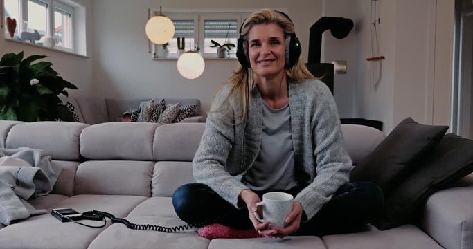 Attractive woman at home sitting cross-legged on the sofa relaxing listening to music on stereo headphones with a smile and cup of coffee in her hands