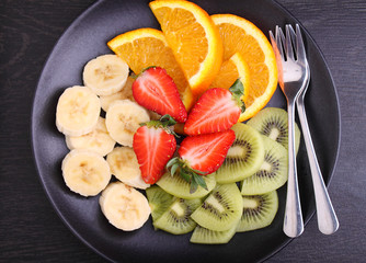 sliced fruit (strawberries, kiwi, orange, banana) on a black plate