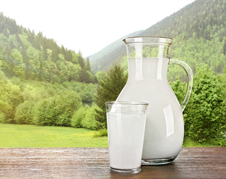 Pitcher, glass of milk on wooden table against forest and mountain background