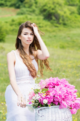Naklejka na ściany i meble Gorgeous young boho fashion woman in white dress on bike with white basket with pink peonies