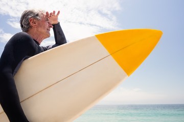 Senior man with surfboard shielding eyes at beach