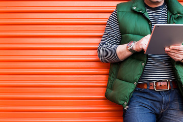 Young man using a tablet in front of an orange garage door, Dressed casually. Jeans, Vest. Urban life style, technology, online, business, shopping, fashion and job hunting concept.