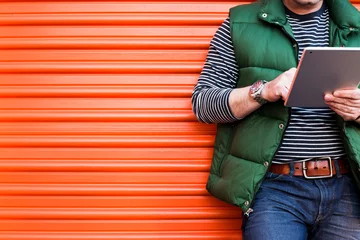 Young man using a tablet in front of an orange garage door, Dressed casually. Jeans, Vest. Urban life style, technology, online, business, shopping, fashion and job hunting concept. © Nicolae Merceanu