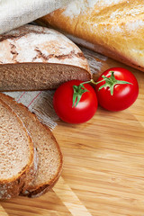 Sliced loaf of gray floured bread, tomatoes and baguette on wooden cutting board