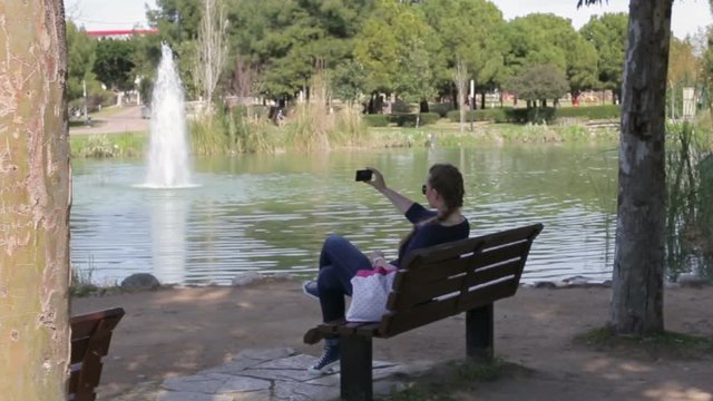 Beautiful happy young girl looking at smartphone screen sitting on a bench in the park in the summer