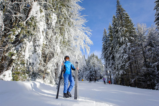 Happy young female skier in winter forest. Woman is wearing blue ski suit helmet and orange glasses, holding skis. Winter sports concept. Carpathian Mountains, Bukovel, Ukraine