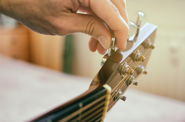 Tuning the guitar. Fingers are turning the tuning peg on the head of acoustic guitar. Authentic shot with blurred room in the background. - obrazy, fototapety, plakaty