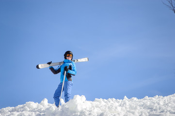 Naklejka na ściany i meble Portrait of young female skier standing on top of the mountain against blue sky on a sunny day. Woman is holding skis on her shoulder smiling and looking into the distance. Winter sports concept.