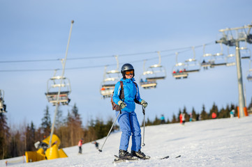 Fototapeta na wymiar Young happy woman skier skiing downhill at ski resort on a sunny day against ski-lift. Girl is wearing blue jacket helmet, goggles and backpack. Carpathian Mountains, Bukovel