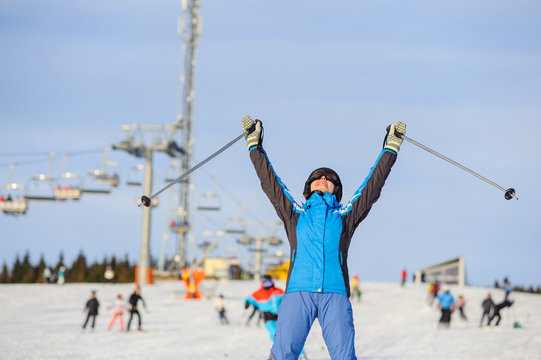 Close-up portrait of happy female skier on a sunny day at ski resort against ski-lift. Woman is standing on a ski slope with raised arms as sign of success. Winter vacation. Bukovel