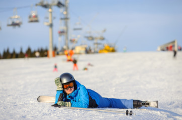 Fototapeta na wymiar Young skier woman in blue ski suit orange goggles and helmet lying on the snow on a sunny day against ski-lift at ski resort. Winter vacation.