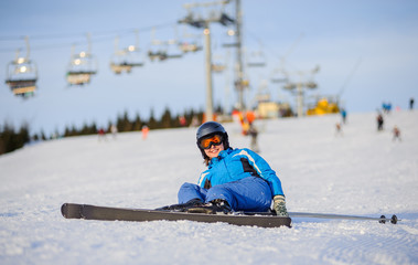 Fototapeta na wymiar Young female skier in blue ski suit smiling after the fall on mountain slope against ski-lift. Ski resort at Carpathian Mountains. Winter sports concept.
