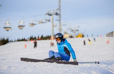 Fototapeta na wymiar Young woman skier in blue ski suit getting up after the fall on mountain slope against ski-lift. Ski resort at Carpathian Mountains, Bukovel. Winter sports concept.