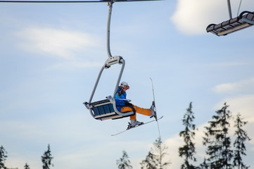 Skier sitting at ski lift against blue sky. Skier is wearing blue jacket, orange pants, helmet and orange goggles. Winter sports concept. Ski resort at Carpathian Mountains, Bukovel, Ukraine