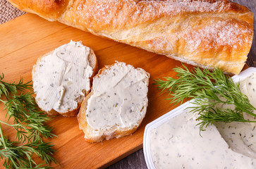 A slice of freshly baked baguette bread and healthy low fat cream cheese with dill and fresh green dill on wooden board.