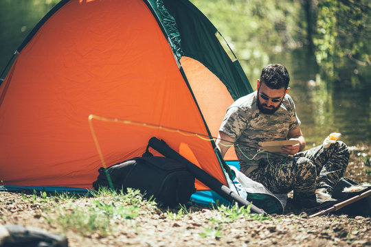 Fly fisherman in camouflage sitting next to the river in his tent and setup  fishing rod. Photos | Adobe Stock