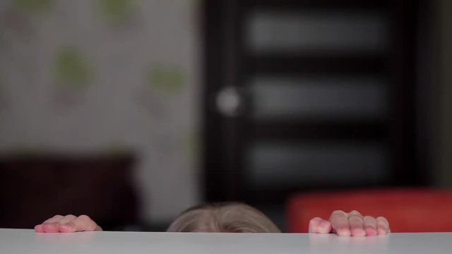Girl coming and looking from under the table then turning eyes to the right and to the left. Then looking straight to the camera and hiding back under the table. 