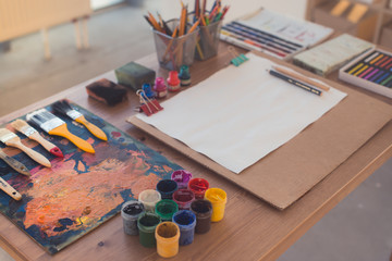 Side view photo of a palette with brushstrokes and paintbrushes. Colorful gouache, crayons sets on the painter workplace.