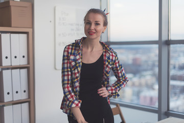 Young woman wearing casual bright shirt standing in light spacious office, her workplace, ready for work day, smiling, looking at camera.