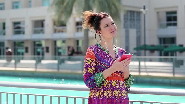 Woman with red smartphone and earphones listens to music. Businesswoman with smartphone. Female with smartphone stands near the water and listens to music. Adult woman uses smartphone with earpieces