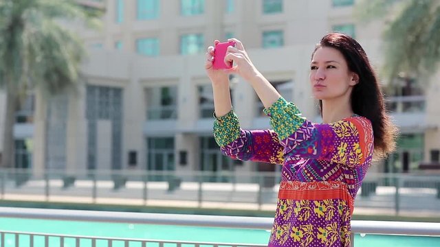 Beautiful woman stands near the blue pool and makes selfie on his smartphone. Female makes photos on his smartphone. Businesswoman with smartphone. Adult woman uses red smartphone, Dubai, UAE