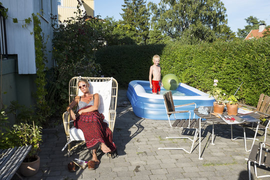 Mother sunbathing with son playing in swimming pool