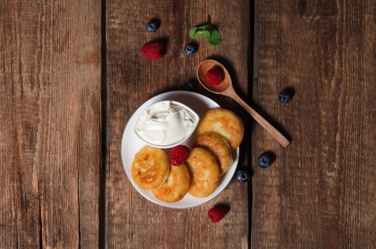 Cottage cheese pancakes with sour cream, raspberries, blueberries and mint, homemade traditional Ukrainian and Russian syrniki on rustic wooden background. Top view.