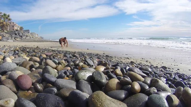Muscular Caucasian man performs bear crawl exercise on pebble stone beach next to surf on sunny day, with native sound