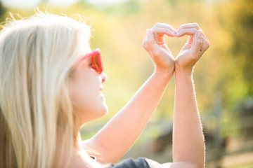 Fototapeta premium Blonde young girl holding hands in heart shape framing. Blonde woman posing outdoor. 