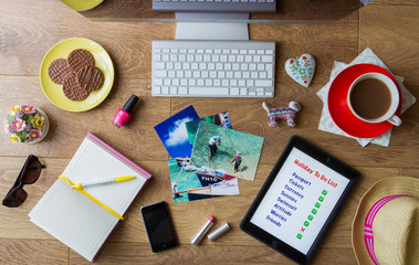 A flat lay image of a desktop full of items needed for booking a vacation online including a tablet with a 'to do list' on, vacation photographs, mobile phone and a cup of coffee.