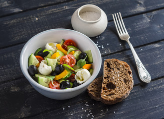 Fresh salad with tomatoes, cucumbers, peppers, olives, cheese  in a ceramic bowl and rye bread ciabatta on dark wooden background