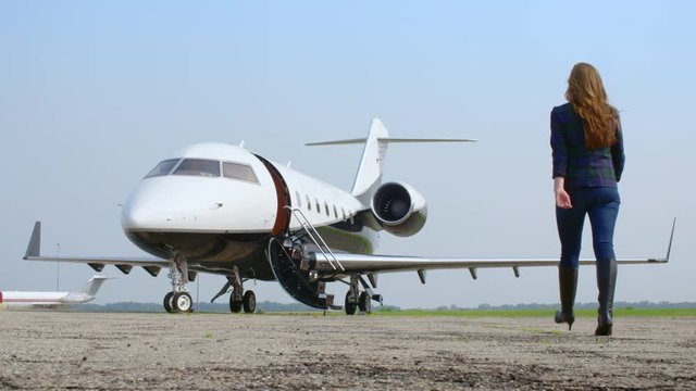 Attractive brunette in a blue jacket and jeans walking towards an executive jet at an airfield.  Wide view, originally recorded in slow motion 4K at 60fps.