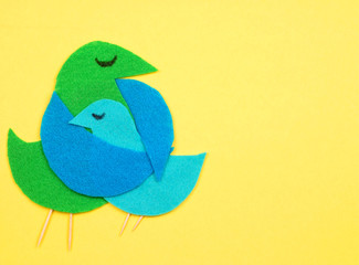 Felt cut out bird shapes in green and blue with toothpick legs and black marker eyes. Mom bird is embracing baby bird. Background is a golden yellow color. Copy space