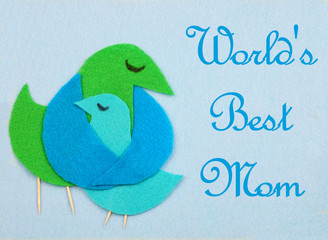 elt cut out bird shapes in green and blue with toothpick legs and black marker eyes. Mom bird is embracing baby bird. Background is baby blue felt. Message added.