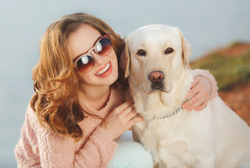 A beautiful young woman,brunette with blue eyes and curly hair,dressed in a white jacket and light trousers, spends time on a rocky beach with his faithful friend, the dog breed white Labrador