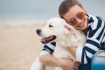Portrait of young beautiful woman in dark glasses,brunette with a beautiful smile, sitting on a sandy beach against the blue of the ocean,embracing a beloved friend - a dog breed Golden Retriever