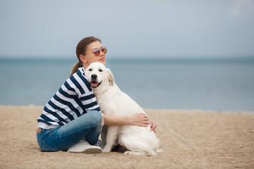 Portrait of young beautiful woman in dark glasses,brunette with a beautiful smile, sitting on a sandy beach against the blue of the ocean,embracing a beloved friend - a dog breed Golden Retriever