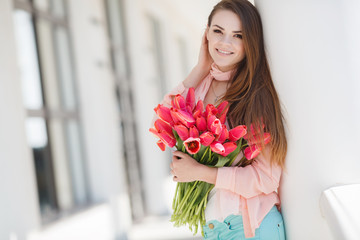 Young beautiful brunette woman with long straight hair and brown eyes,dressed in a beige jacket and a white t-shirt posing in summer on the street with a bouquet of red tulips